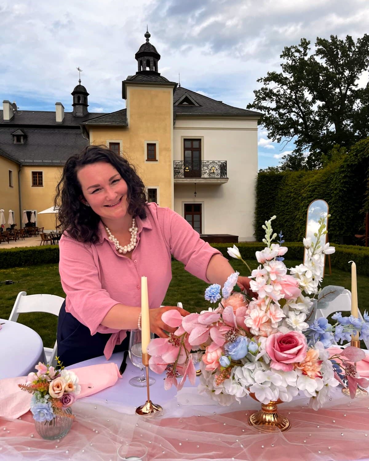 Zuzana Golcová s úsměvem aranžuje květinovou výzdobu slavnostní tabule v zámecké zahradě